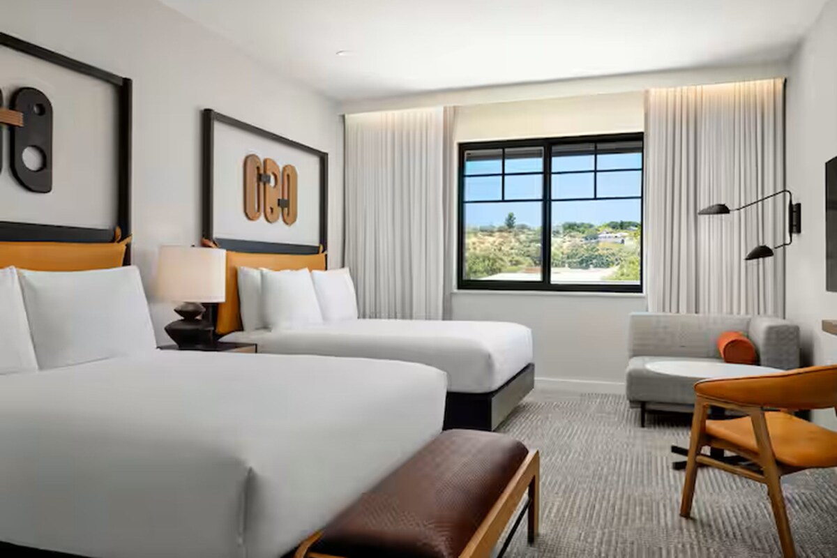 A hotel room with two beds, a sofa, a chair, a window with a view, and artwork on the walls.