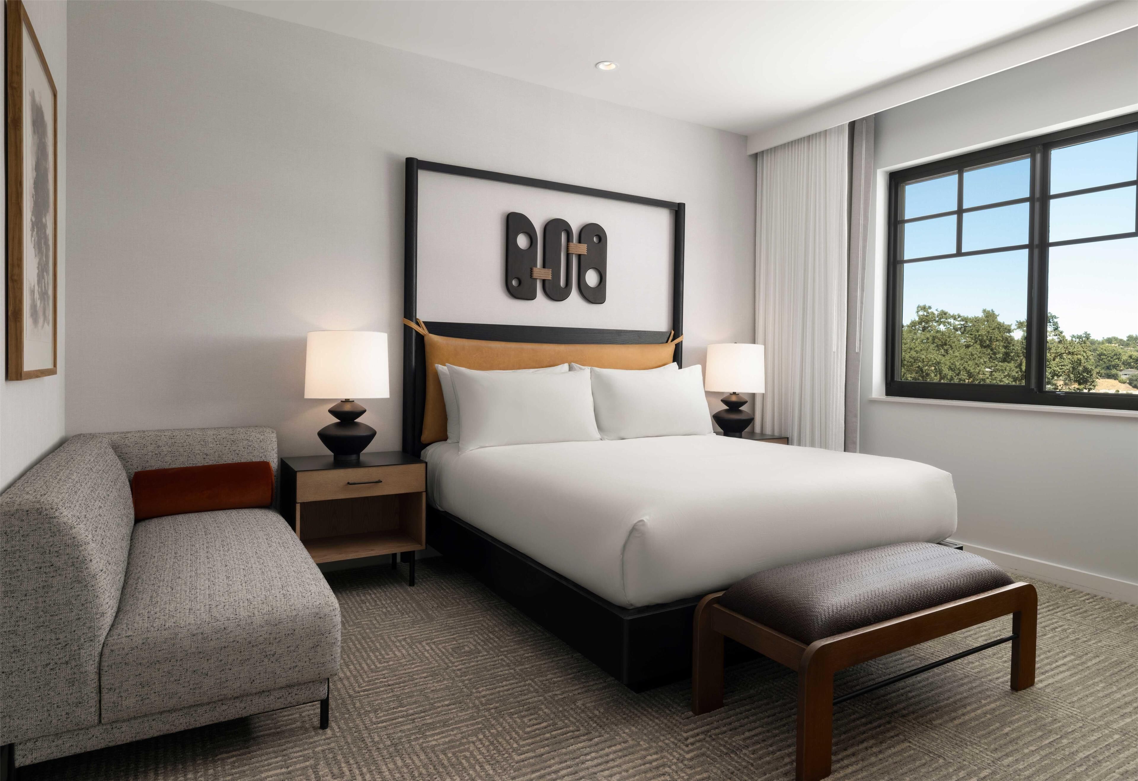 Habitación de hotel moderna con una cama grande, una mesita de noche con lámparas, una silla y una ventana con vistas a árboles.