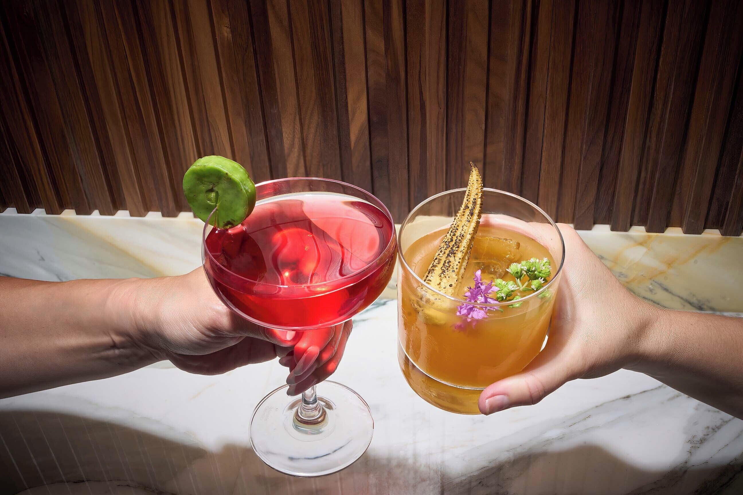 Two hands holding cocktail glasses with garnishes, one with a lime wedge and the other with a decorative item and a flower.