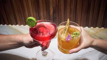 Two hands holding cocktail glasses with garnishes, one with a lime wedge and the other with a decorative item and a flower.
