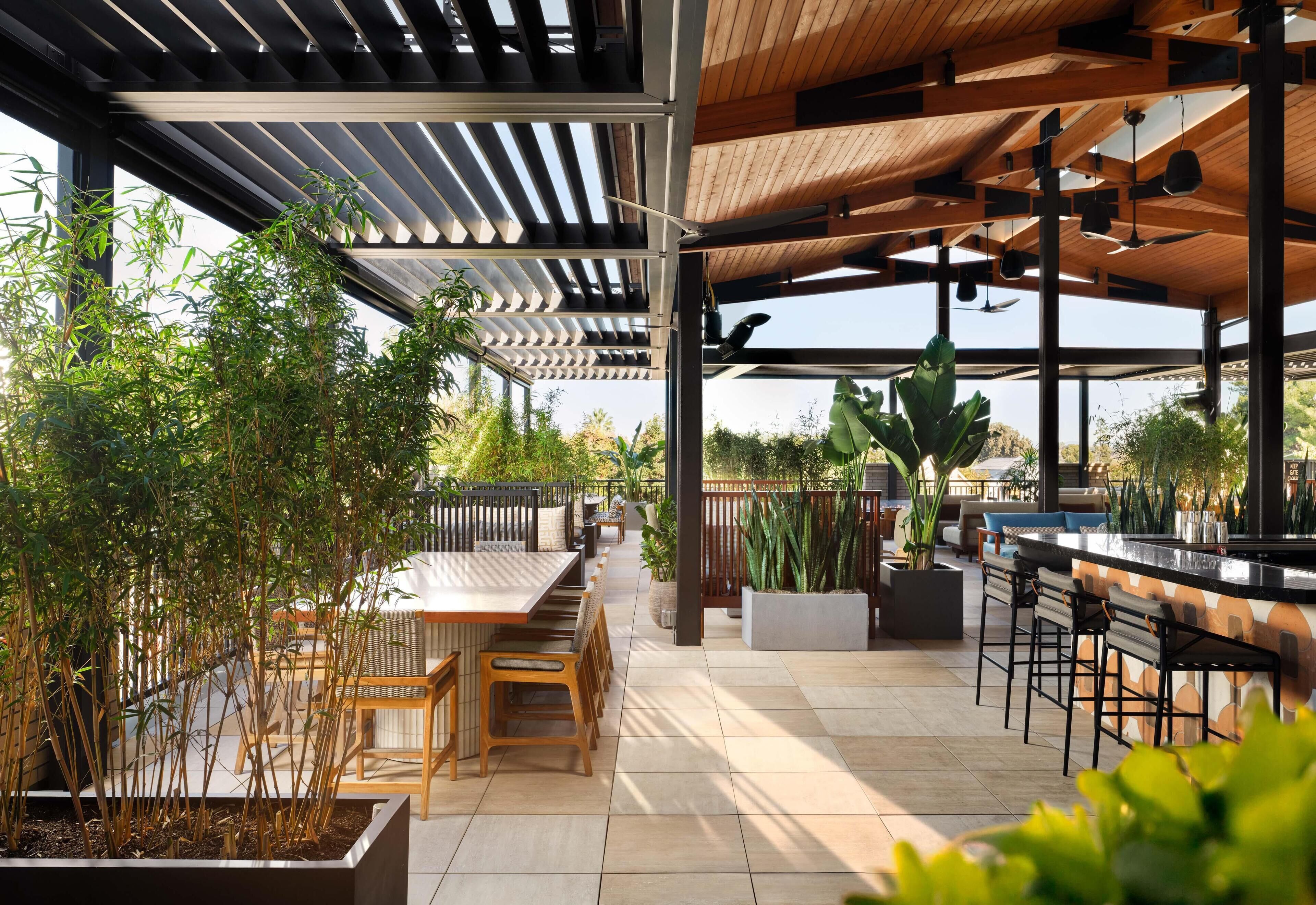 An outdoor patio area with wooden tables and chairs, potted plants, and a bar counter.