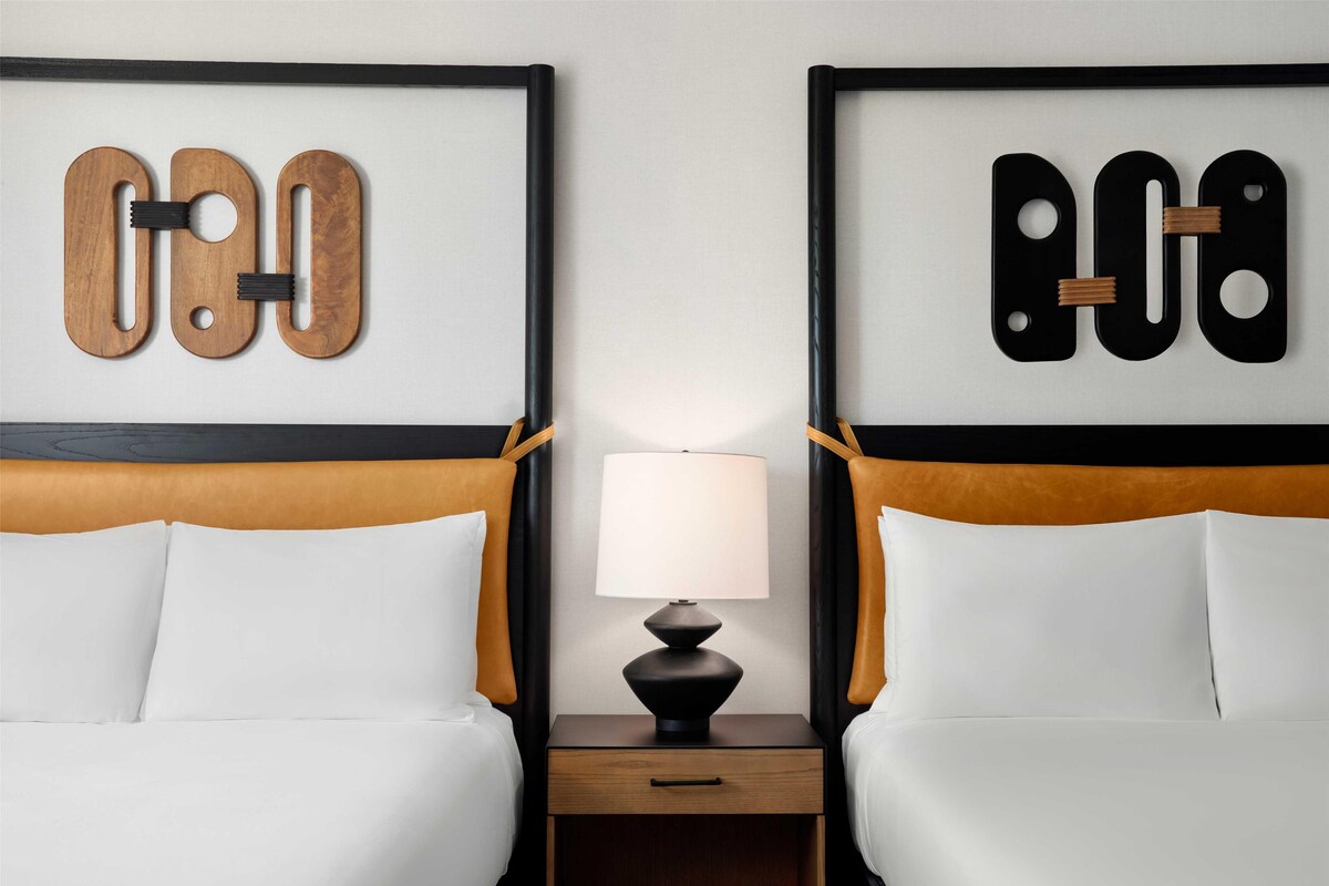 A hotel room with two beds, a nightstand with a lamp, and artwork on the headboards.