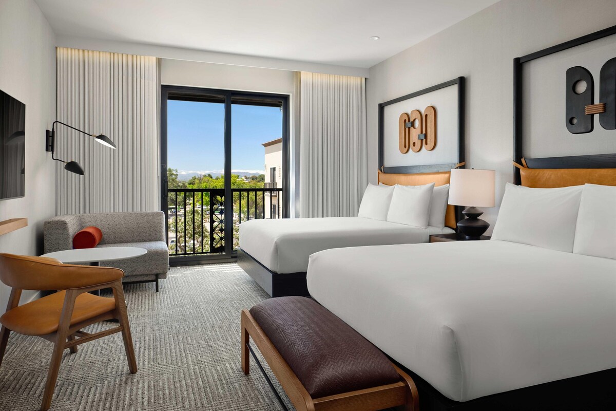 A hotel room with two beds, a sofa, a chair, a bench, a TV, and a balcony with a view.