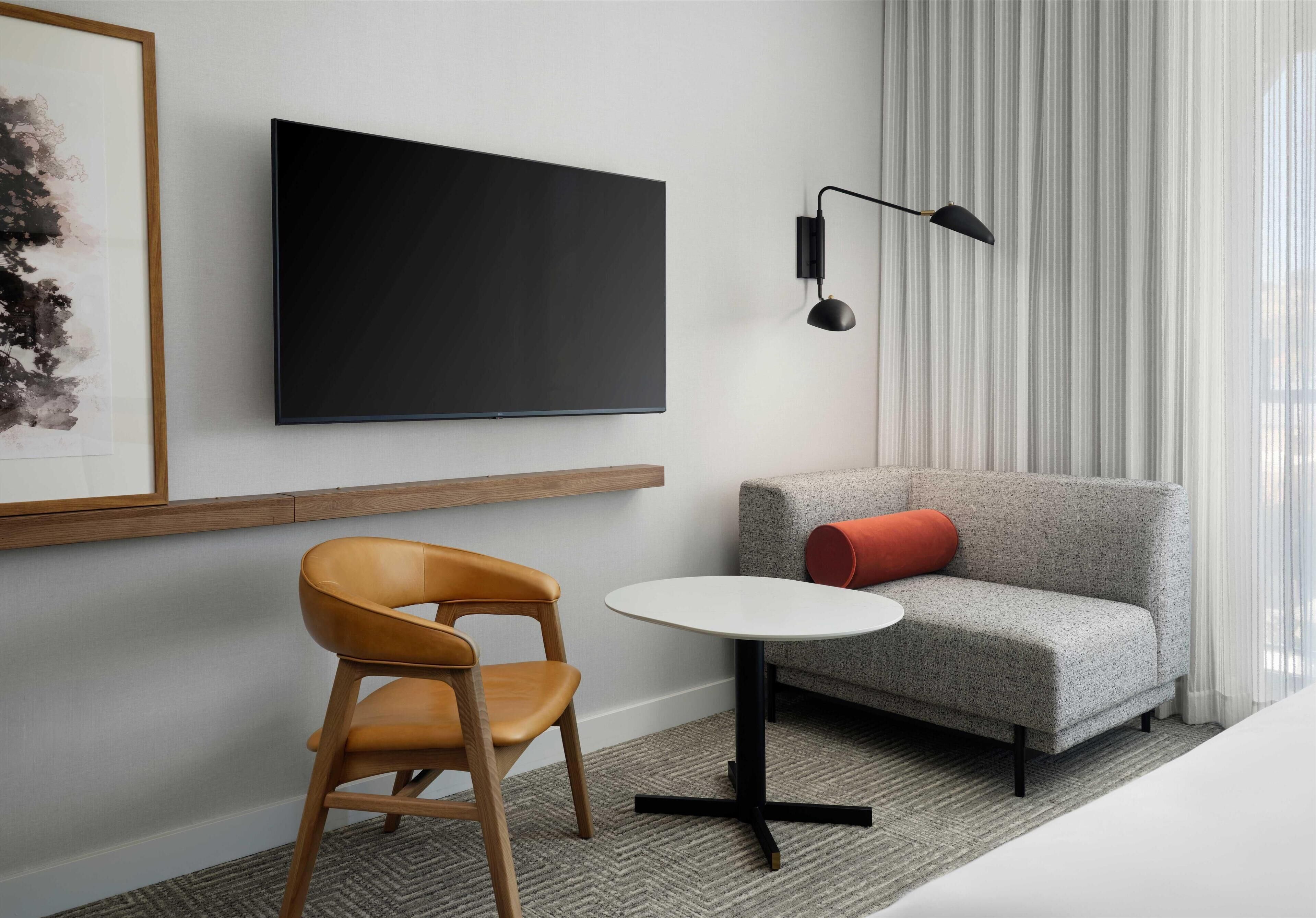 Une chambre d’hôtel moderne équipée d’un téléviseur à écran plat fixé au mur, d’une petite table ronde, d’une chaise en bois, d’un canapé gris agrémenté d’un coussin rouge et d’un lampadaire noir.