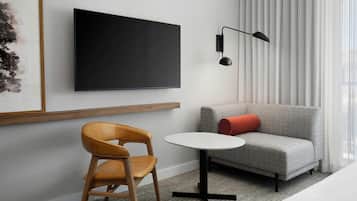 Une chambre d’hôtel moderne équipée d’un téléviseur à écran plat fixé au mur, d’une petite table ronde, d’une chaise en bois, d’un canapé gris agrémenté d’un coussin rouge et d’un lampadaire noir.