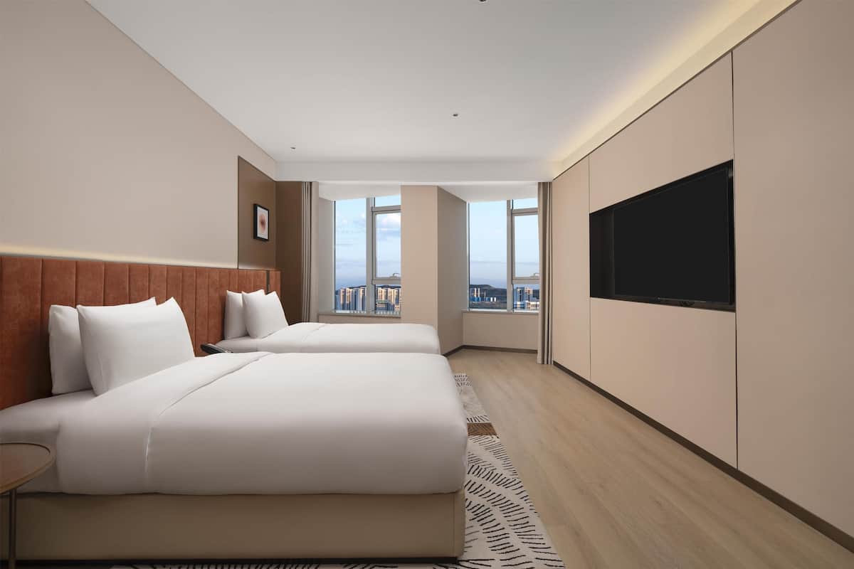 A modern hotel room with two beds, a large TV, and a city view.