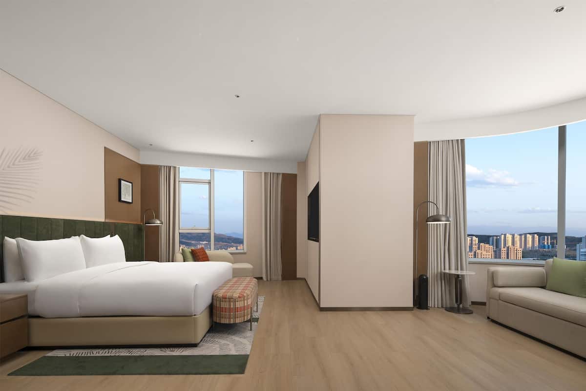 A modern hotel room with a large bed, a sofa, a small table, and a TV. The room has a city view.