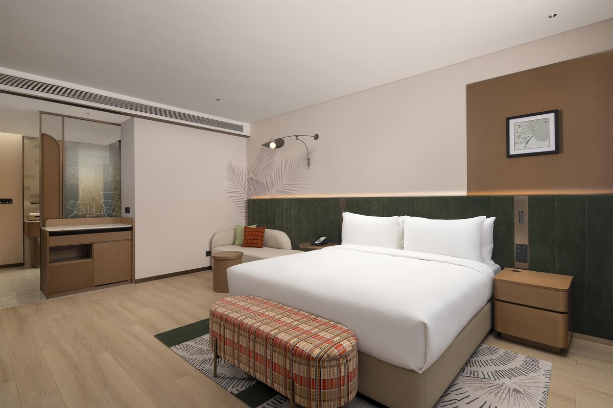 A modern hotel room with a large bed, a desk, a chair, a small table, a telephone, and a framed picture on the wall.