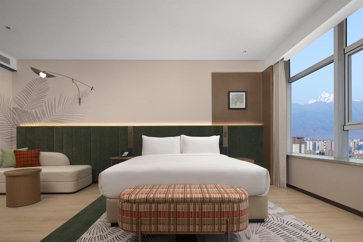 A modern hotel room with a large bed, a small sofa, a side table, a lamp, and a view of a cityscape through a large window.