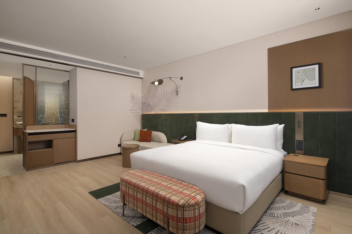 A modern hotel room with a large bed, a desk, a chair, a bench, a mirror, and a nightstand.