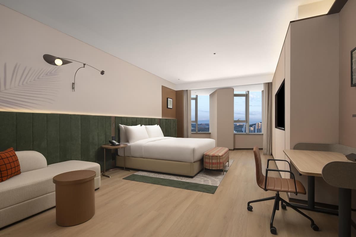 A modern hotel room with a bed, a desk, a chair, a small table, and a view of the city.