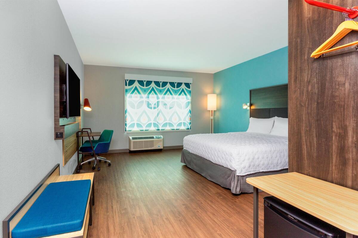 A hotel room with a bed, a desk, a chair, a TV, and a window with patterned curtains.