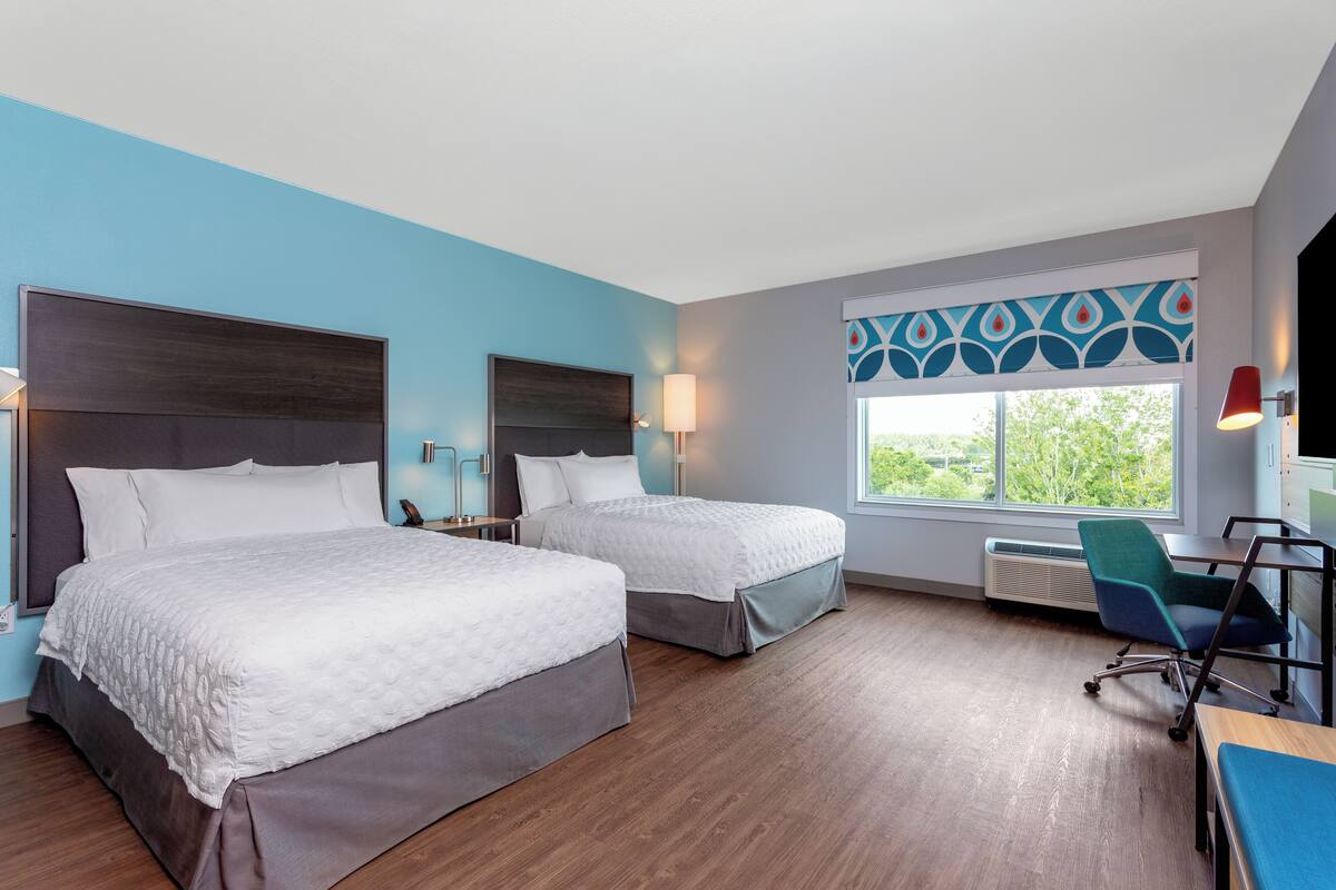 A hotel room with two beds, a desk, a chair, a window with a view, and a patterned curtain.