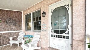 A porch with two Adirondack chairs, a decorative fish wall, and a large window with a welcome sign. - Coastal Cottage 225 - Walk to Beach, King Bed (Cocoa Beach)
