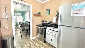A compact kitchen with a refrigerator, stove, microwave, and a dining area with chairs and a table. - Coastal Cottage 225 - Walk to Beach, King Bed (Cocoa Beach)