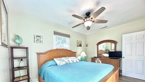 A bedroom with a wooden bed, a ceiling fan, a nightstand, a lamp, a dresser, a television, and a window with a view of trees. - Coastal Cottage 225 - Walk to Beach, King Bed (Cocoa Beach)