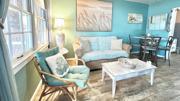 A living room with a teal wall, a wooden floor, a sofa, a rocking chair, a coffee table, and a ceiling fan. - Coastal Cottage 225 - Walk to Beach, King Bed (Cocoa Beach)