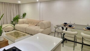 A modern living room with a beige sofa, a glass coffee table, and a dining area with a table set for two.