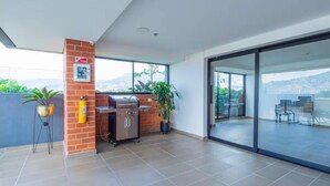 A modern outdoor patio with a barbecue grill, potted plants, and a glass door leading to a balcony with a city view.