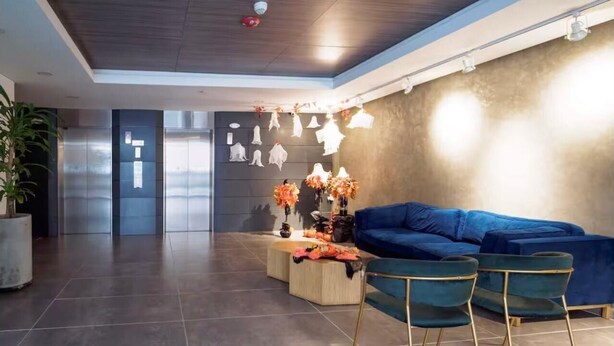 A modern lobby with a blue sectional sofa, two green chairs, a wooden coffee table, and a wall decorated with hanging white figures and orange flowers.