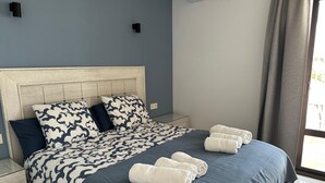 A neatly made bed with a patterned bedspread, two pillows, and rolled towels on a clean, tiled floor.