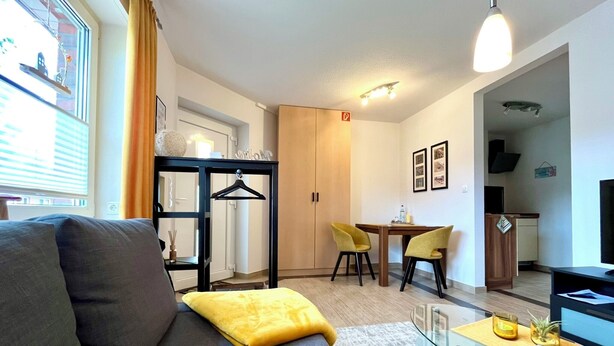 Un salon moderne avec un canapé gris, un coussin jaune, une table basse en verre et un coin repas comprenant deux chaises jaunes.