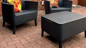 Une terrasse avec deux canapés en osier et une table, un mur en briques et une fenêtre avec des stores.