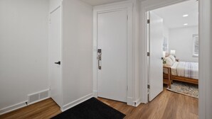 Un pasillo con paredes blancas y piso de madera que conduce a un dormitorio con cama, mesita de noche y lámpara.