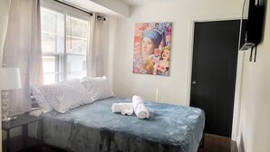 Une chambre à coucher avec un lit, des oreillers, une table de chevet avec une lampe, une fenêtre avec des stores et une porte.