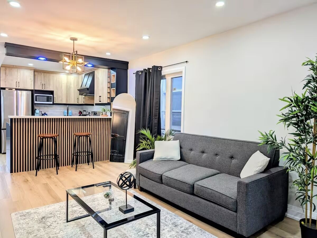 Ein modernes Wohnzimmer mit grauem Sofa, einem Glastisch und einer Küchenzeile mit Barhocker und Mikrowelle.