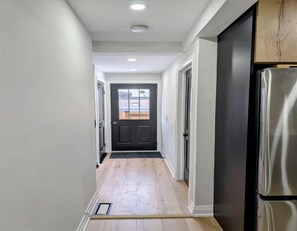 A hallway with a door leading to a room, a refrigerator, and a window with blinds. - Bright 2BR Home – Walk to Shops & Restaurants (Hamilton)