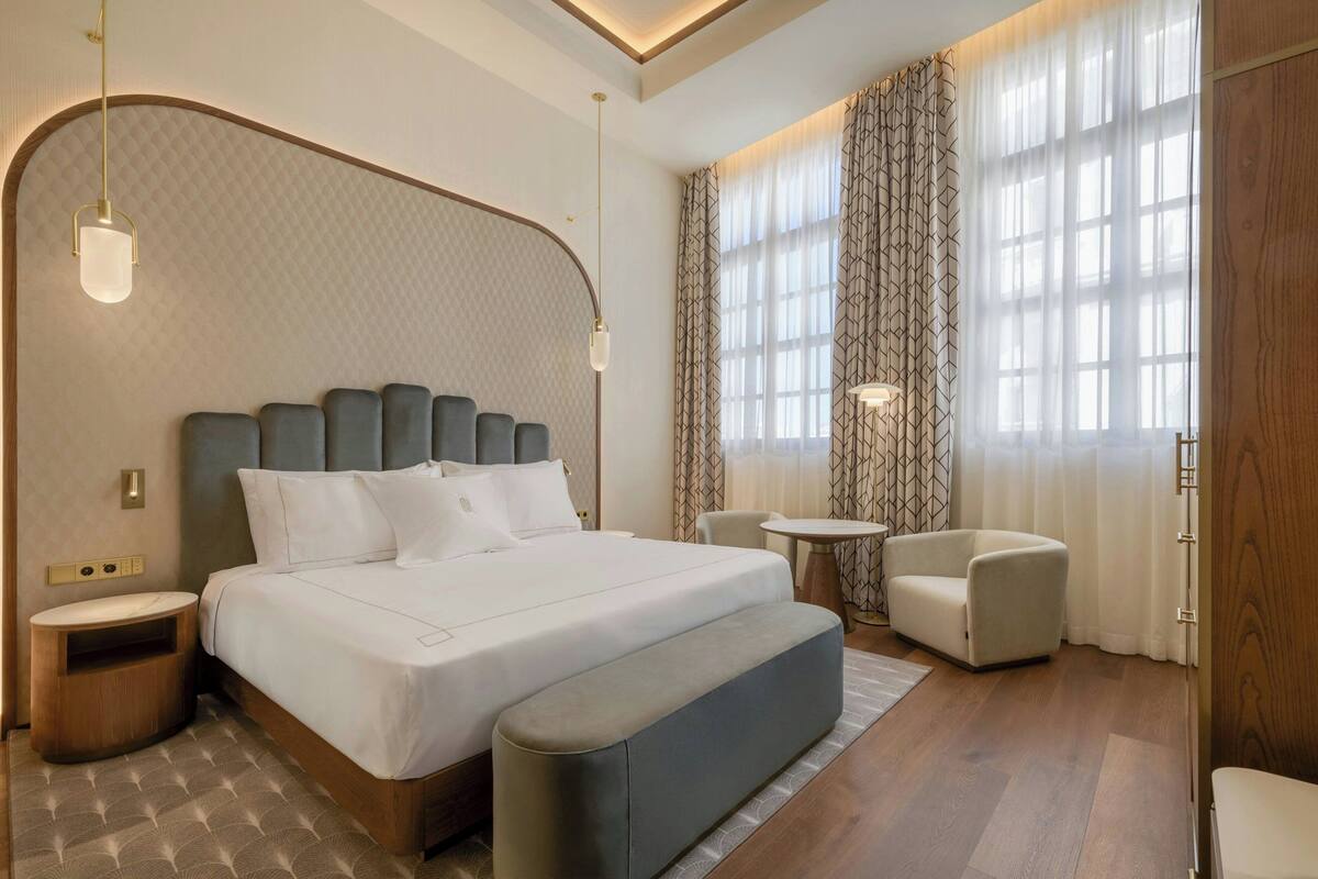 A modern hotel room with a large bed, a small round table, a sofa, and a large window with curtains.