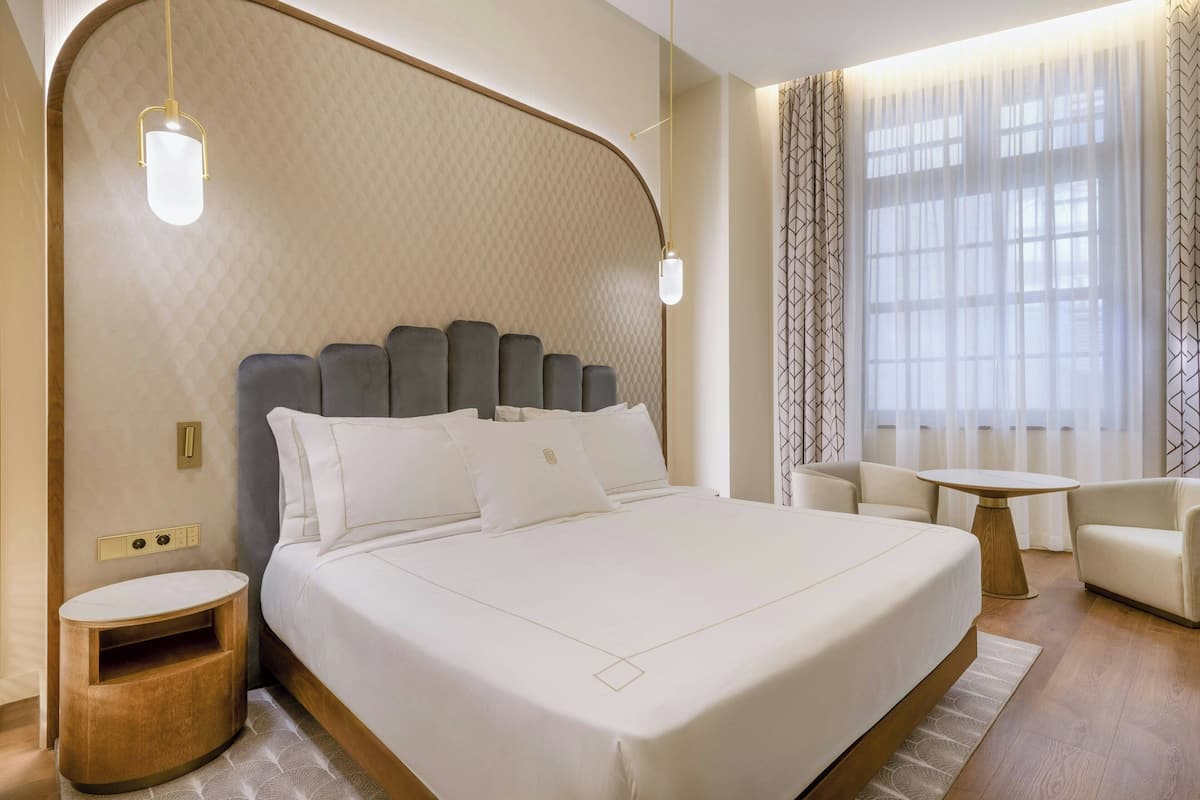 A modern hotel room with a large bed, a bedside table, a small round table, and a sofa.