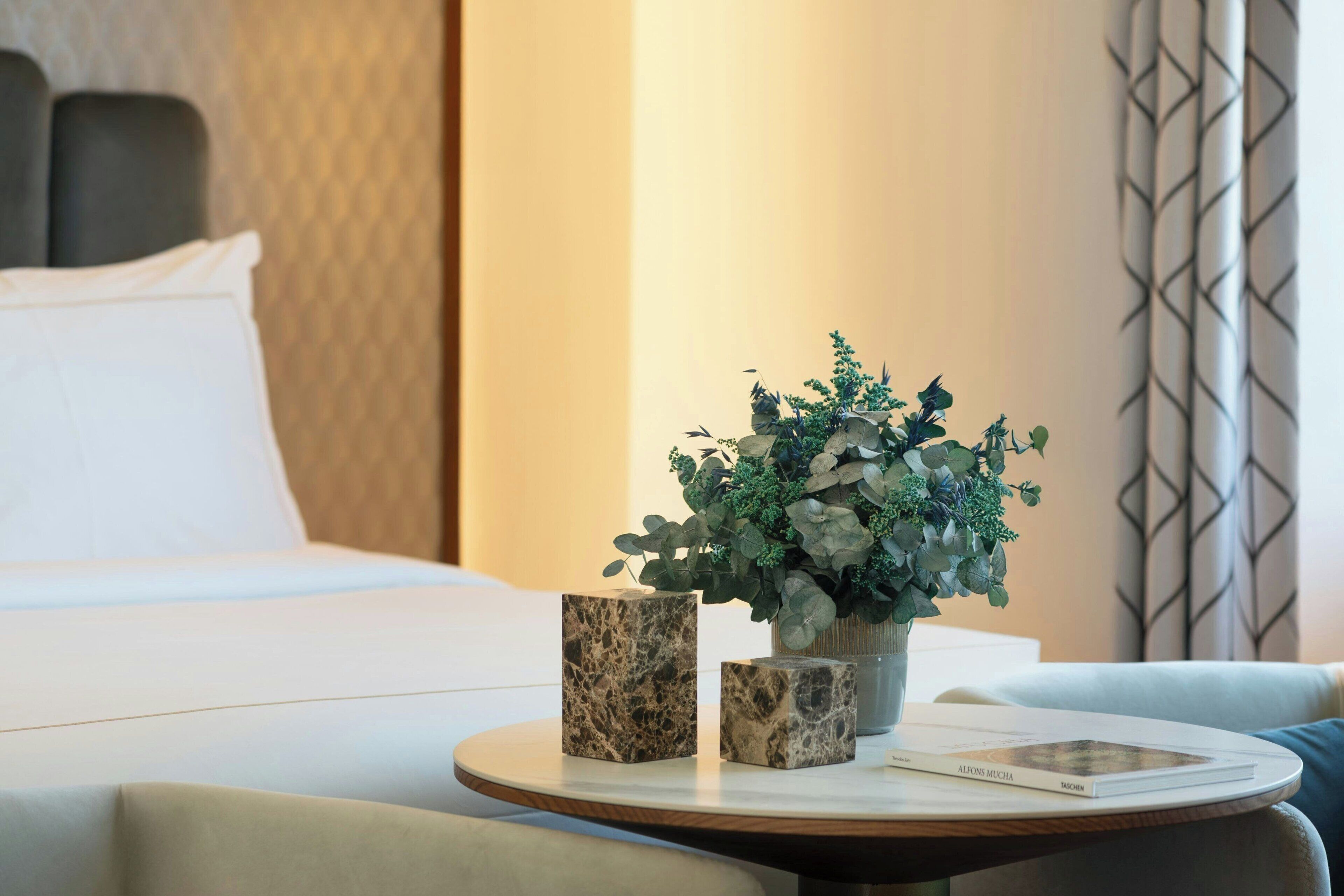 A hotel room with a bed, a round table with a vase of green plants, two marble cubes, and a book titled "ALFONS MUCHA".