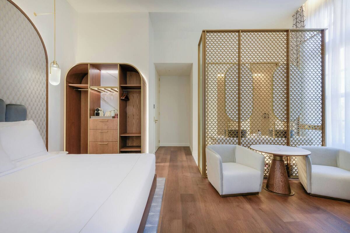 A hotel room with a bed, a wooden wardrobe, a seating area with two chairs and a table, and a large window.