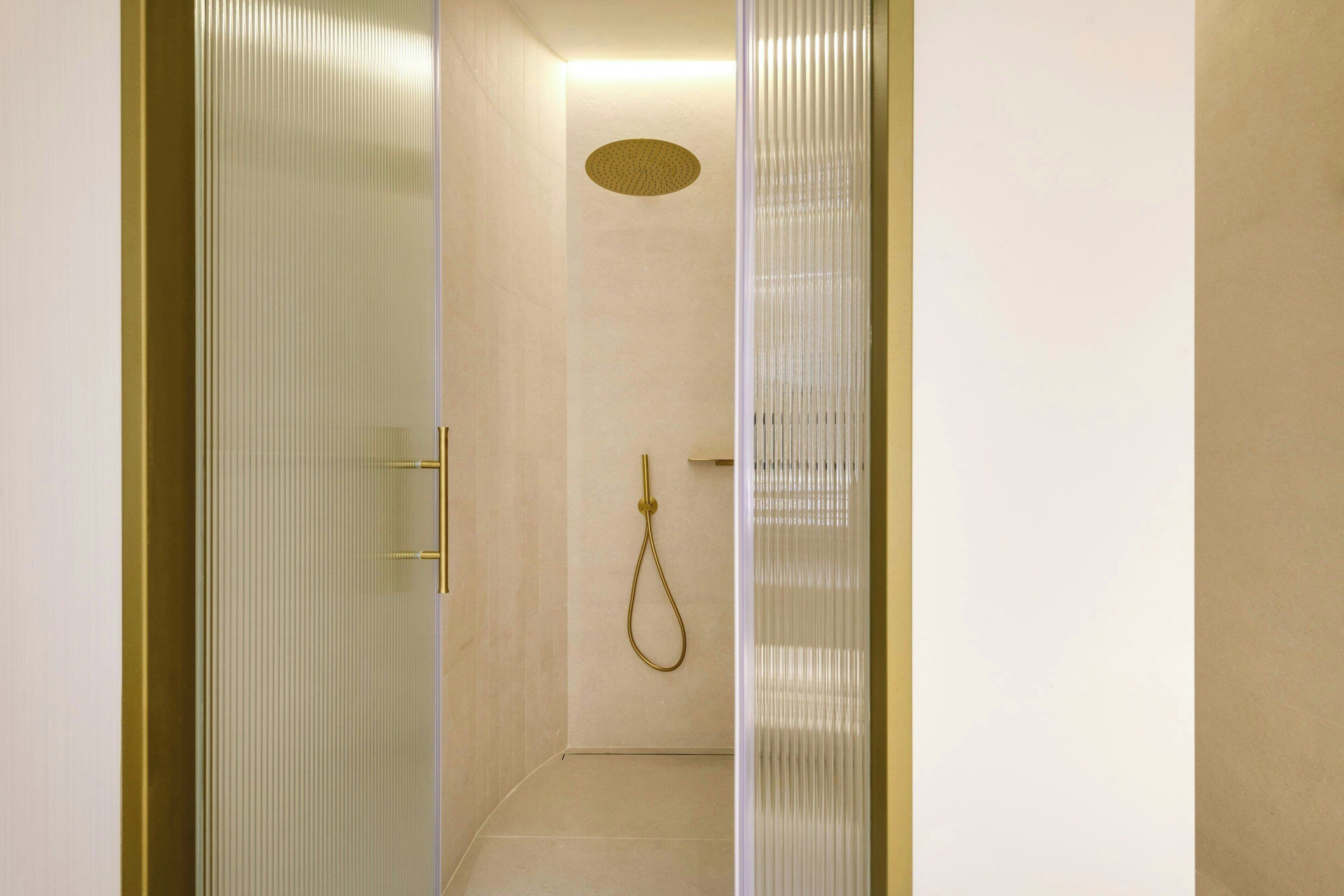 A narrow, glass-walled walk-in shower with a gold showerhead and a ceiling-mounted circular light fixture.