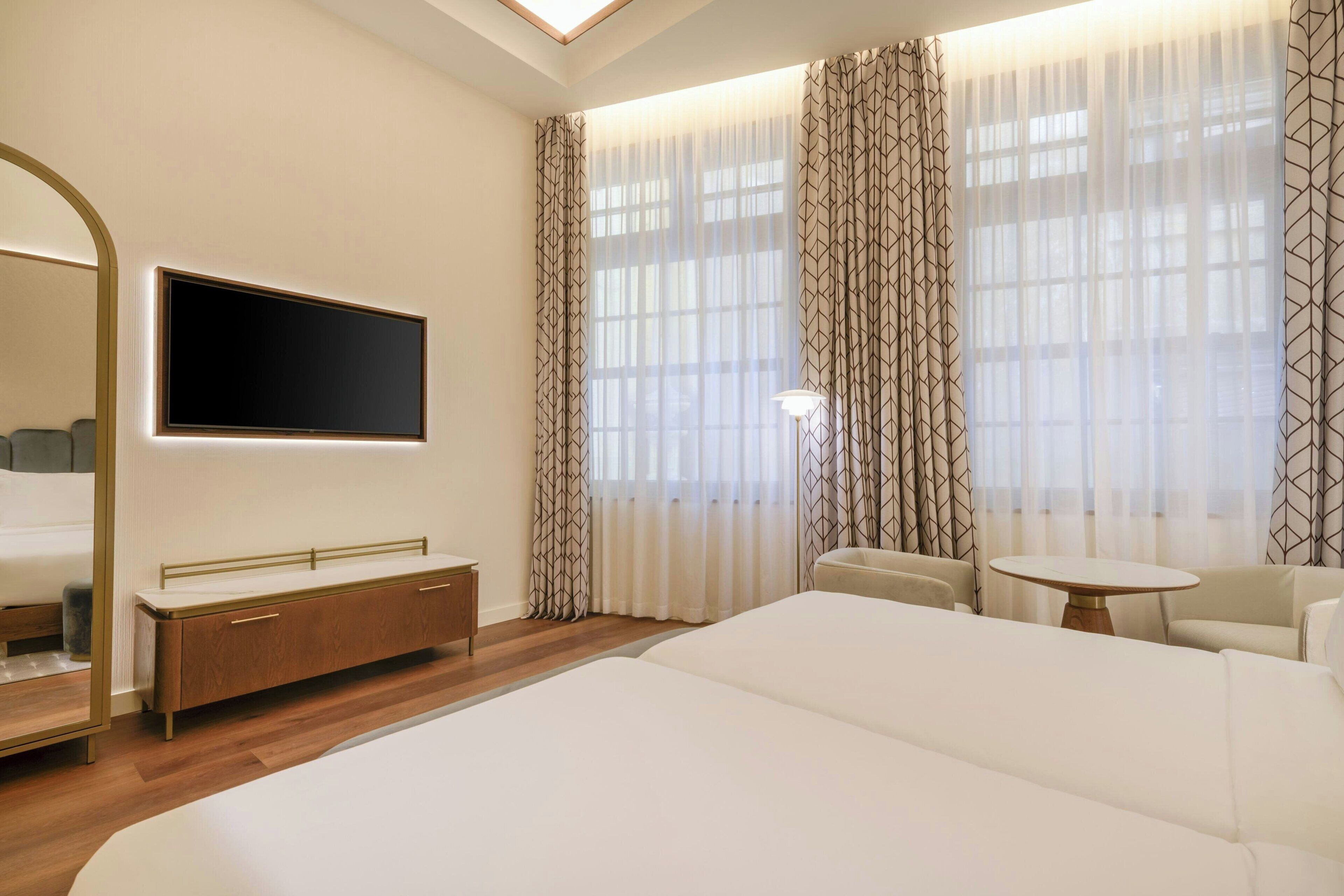 A modern hotel room with a large bed, a TV mounted on the wall, a wooden bedside table, a small round table, and a sofa.