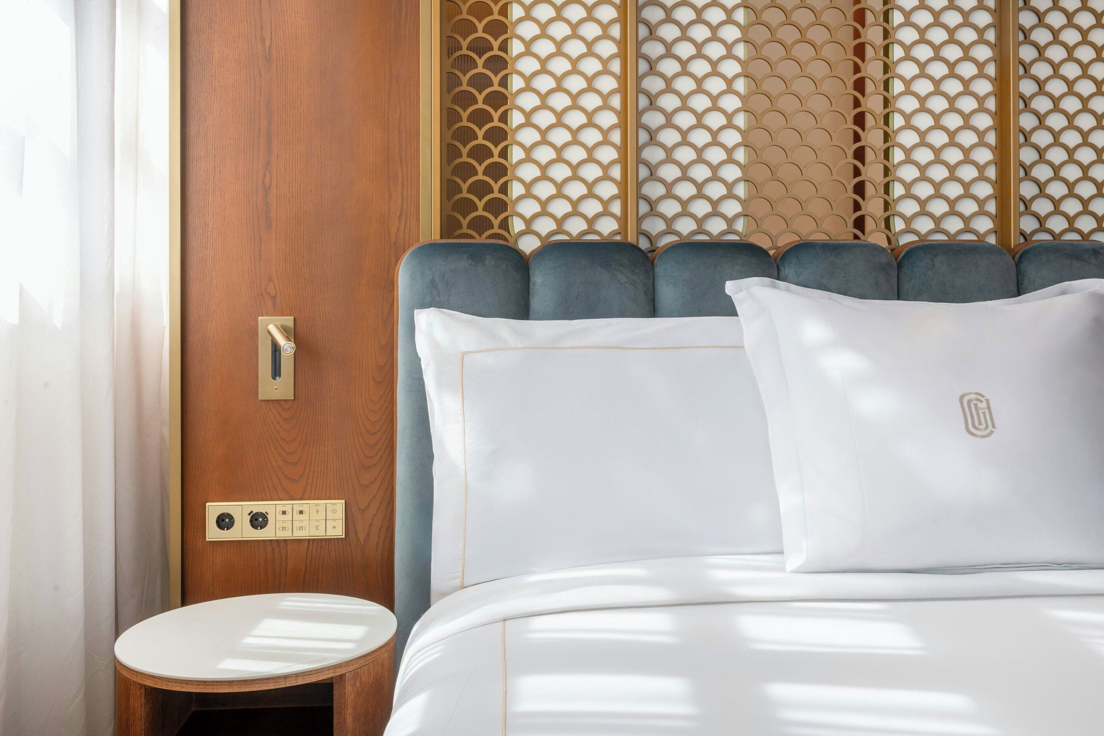 Habitación de hotel con puerta de madera, cama con ropa blanca, una pequeña mesa redonda y un panel decorativo en la pared.