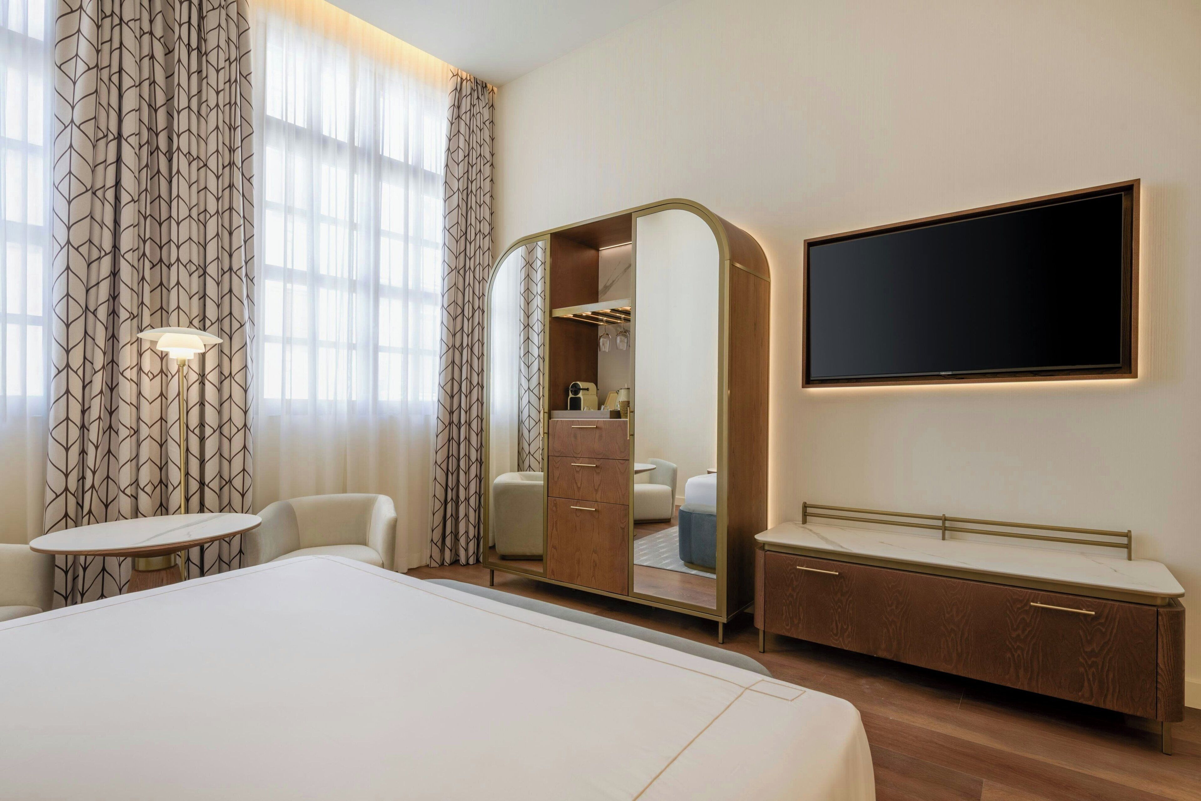 A hotel room with a large bed, a bedside table, a mirror cabinet, a TV, and a window with curtains.