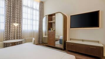A hotel room with a large bed, a bedside table, a mirror cabinet, a TV, and a window with curtains.