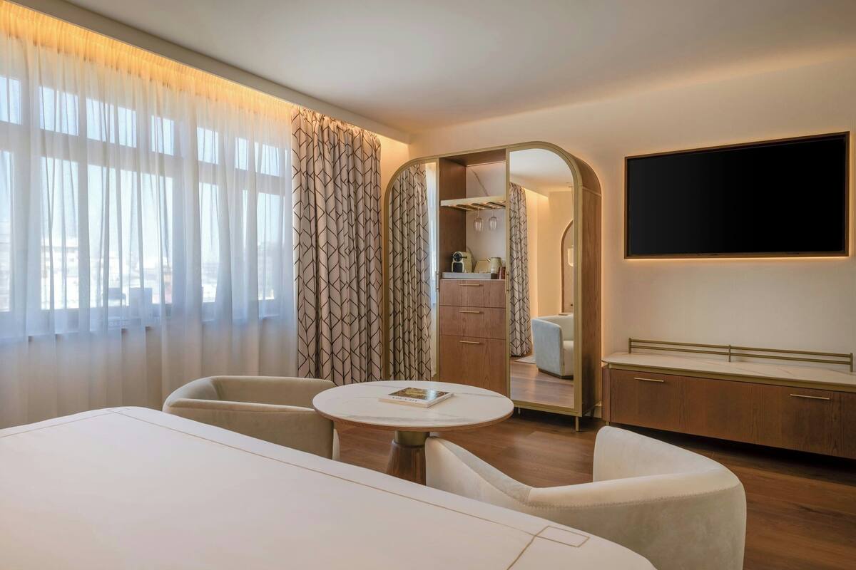 A hotel room with a large bed, a round table with chairs, a TV mounted on the wall, and a view of the city through sheer curtains.