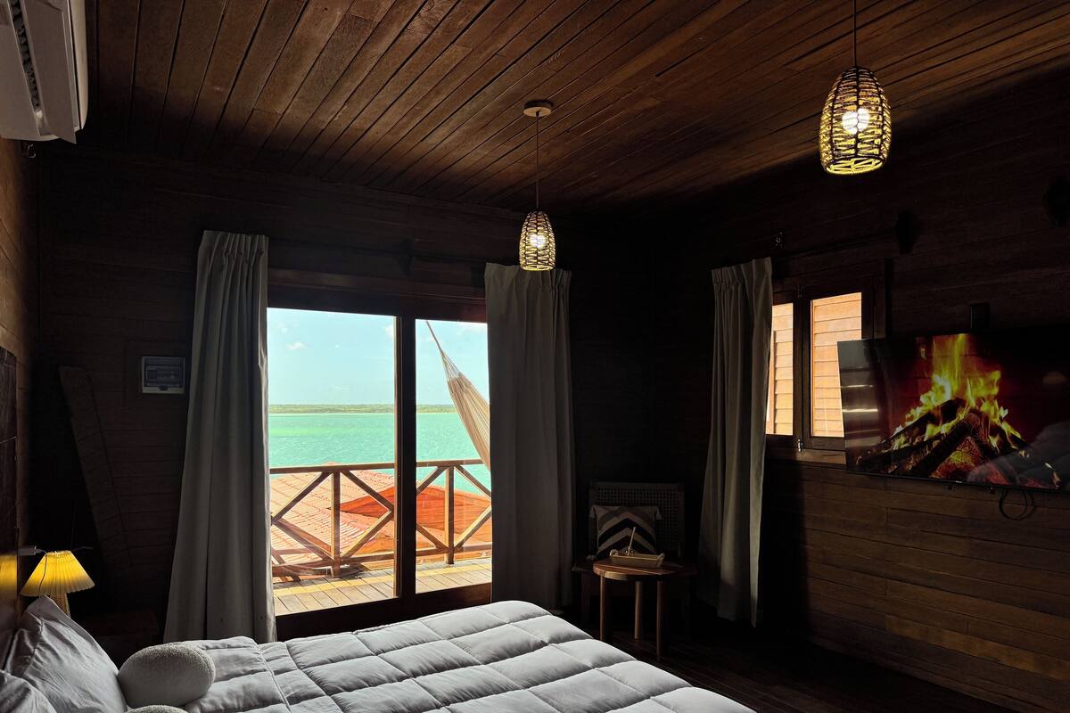 A room with a bed, a TV displaying a fire, and a view of the ocean through a window.