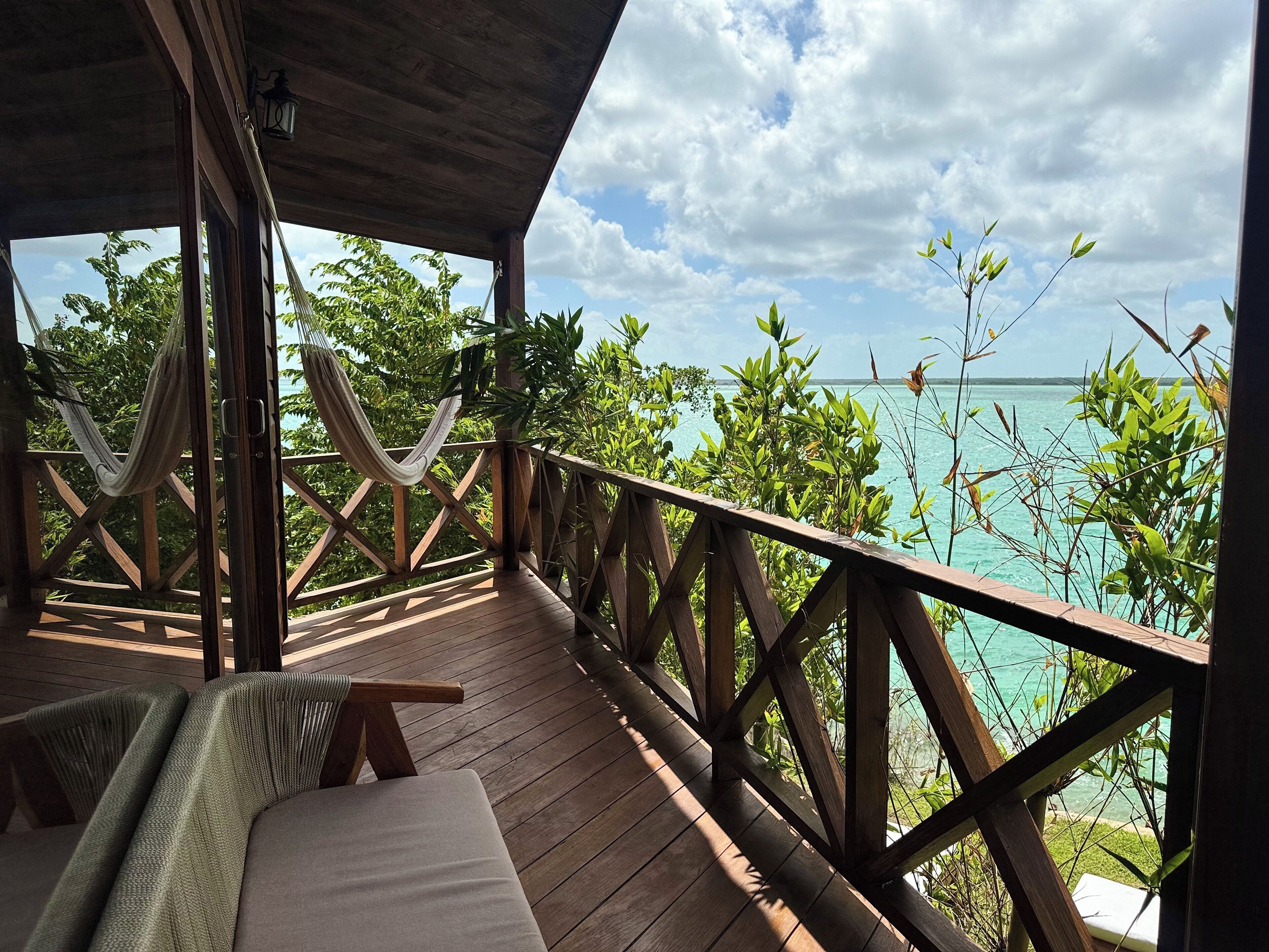 A wooden deck with a hammock, a cushioned bench, and a view of turquoise waters and greenery.
