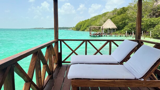 Une terrasse en bois avec une balustrade en bois, deux chaises longues blanches avec coussins, et une vue sur des eaux turquoise et une structure à toit de chaume en arrière-plan.