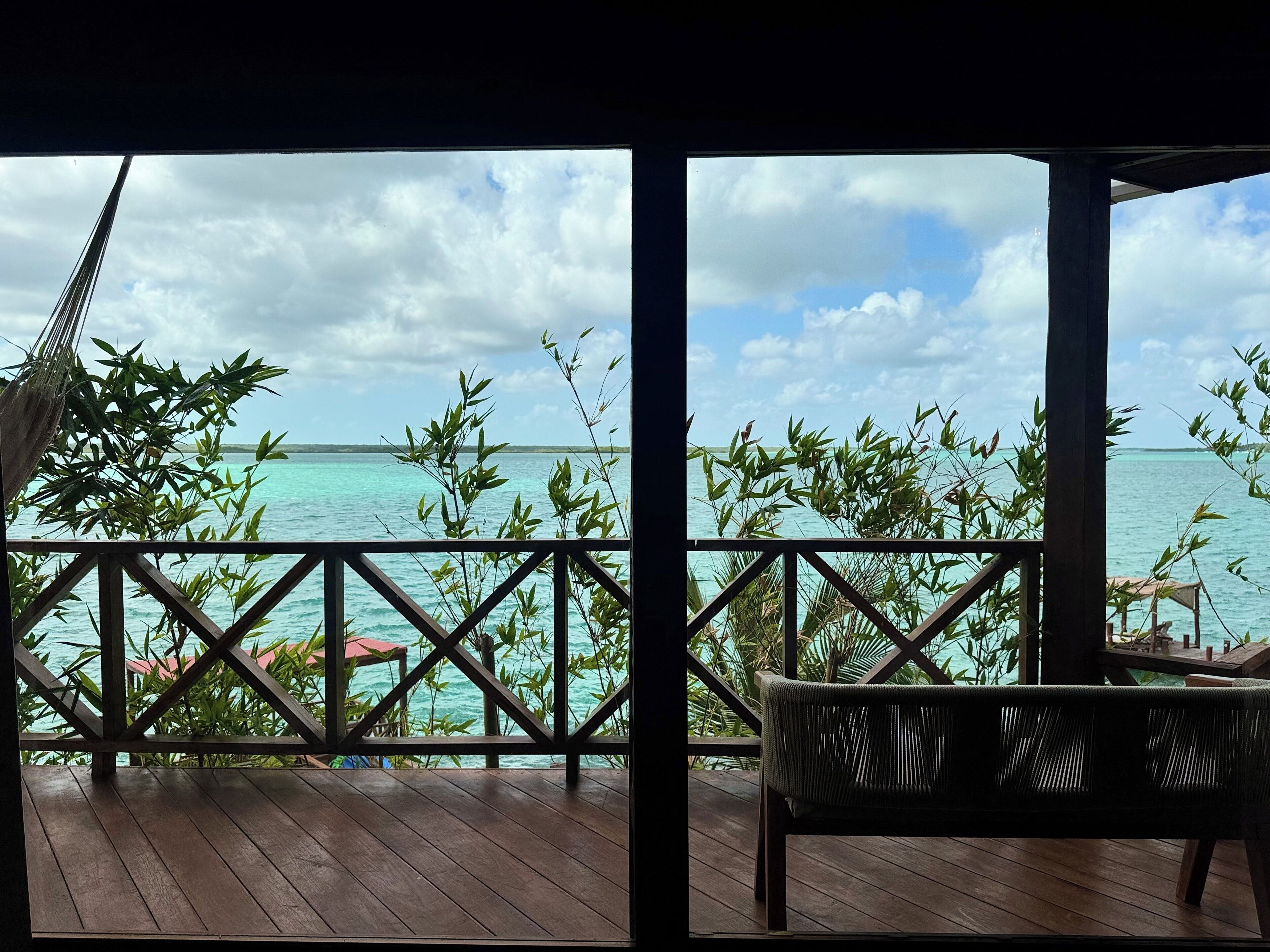 A wooden deck with a view of turquoise waters and distant islands, bamboo plants, and a hammock.