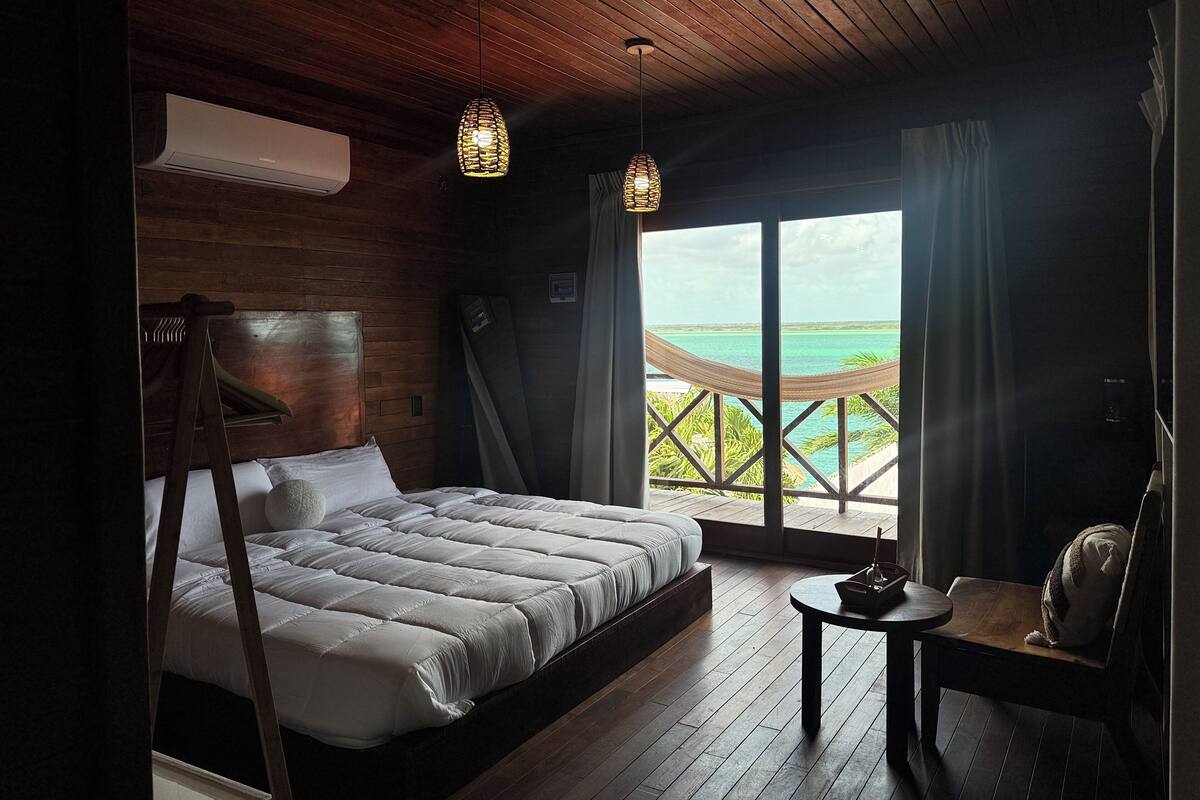 A bedroom with a large bed, a wooden ceiling, and a view of the ocean through a large window.