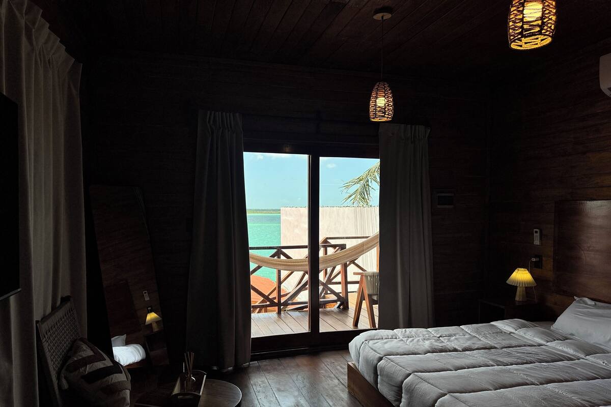 A bedroom with a large bed, a small table with a lamp, and a view of the ocean through a sliding door.