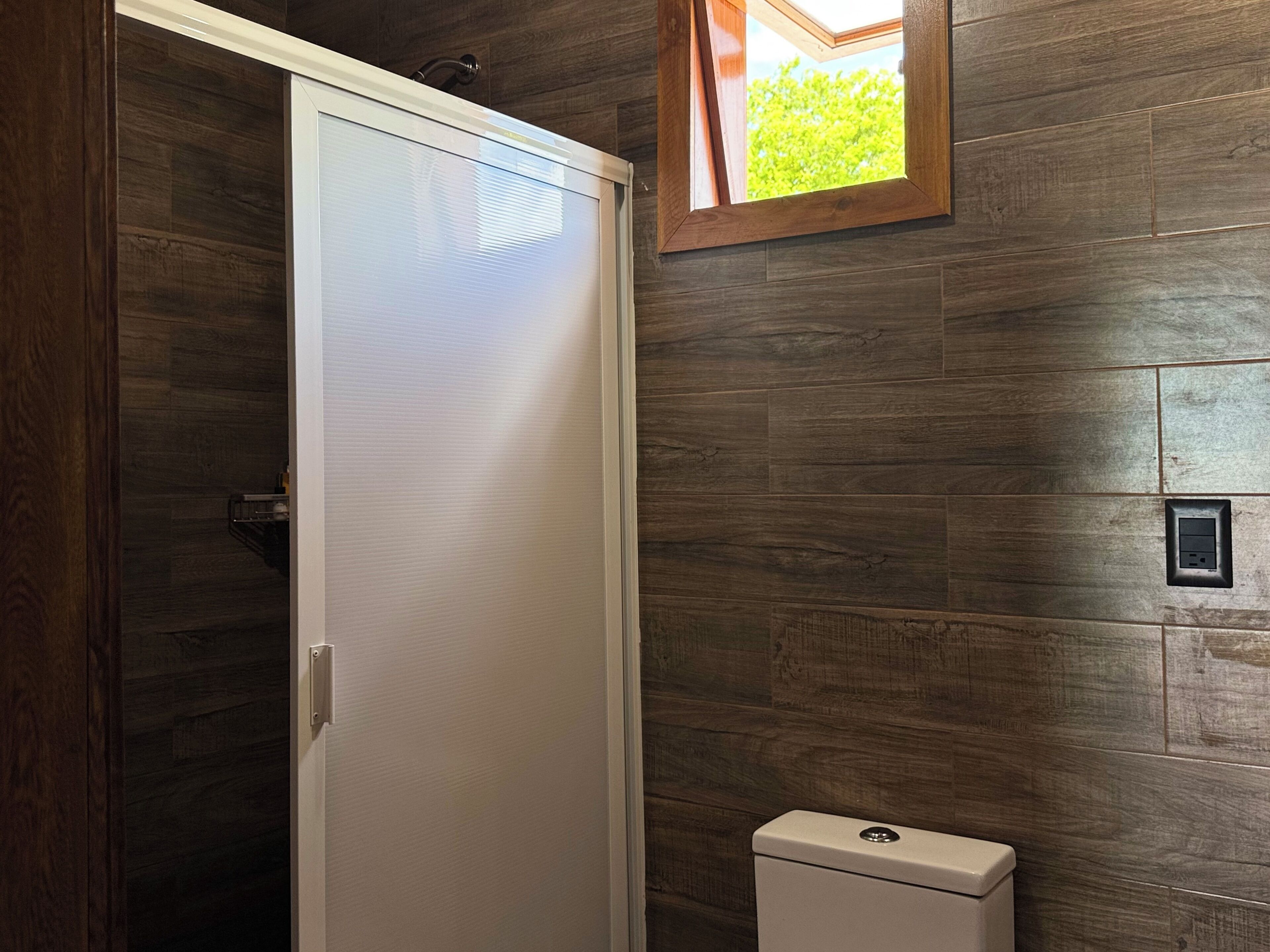 A bathroom with a wooden wall, a white toilet, and a window with a view of greenery.