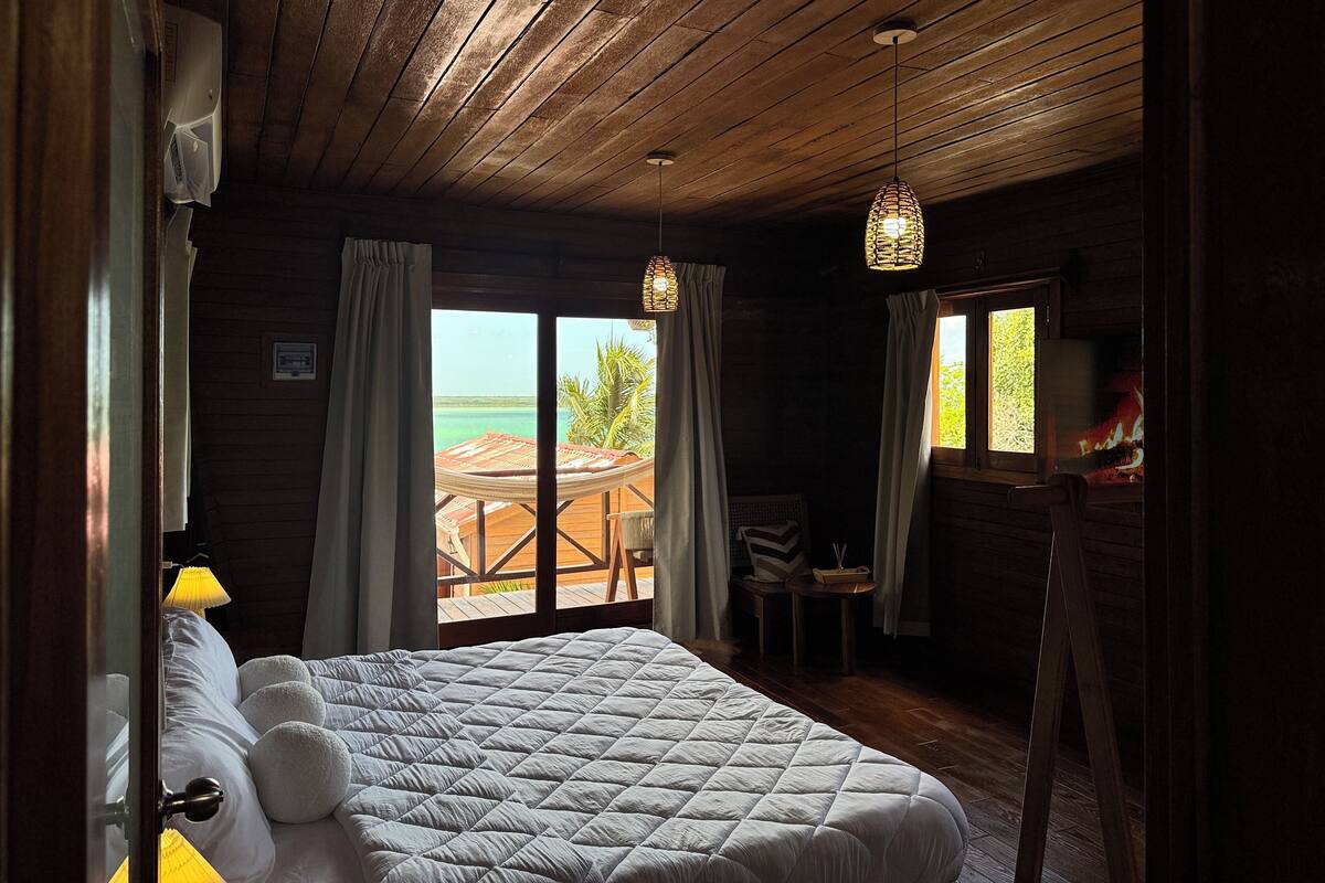A wooden cabin room with a bed, a small table, and a view of the ocean.