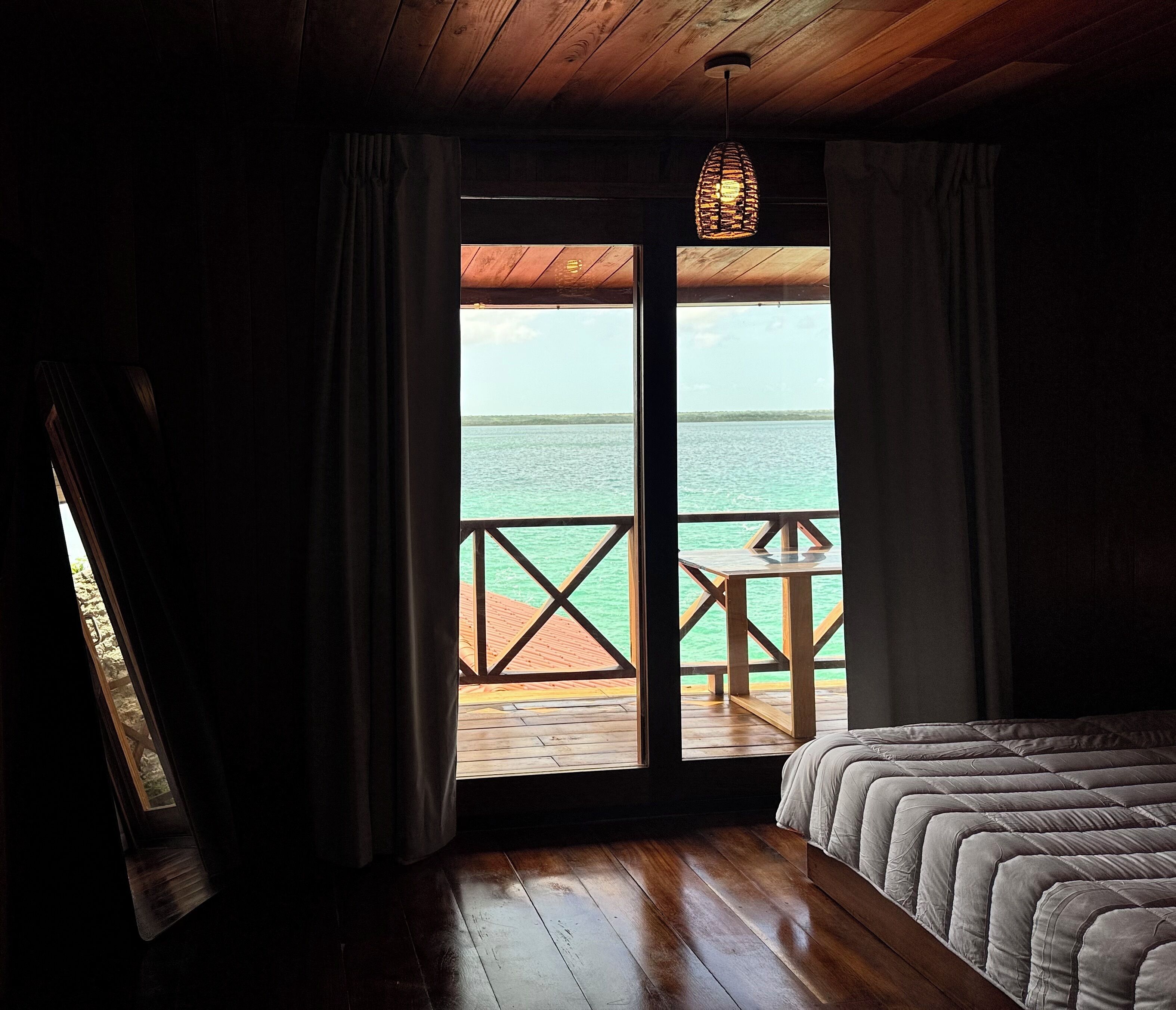Een slaapkamer met een houten vloer, een bed met een grijze dekbedovertrek en uitzicht op zee door een glazen deur.